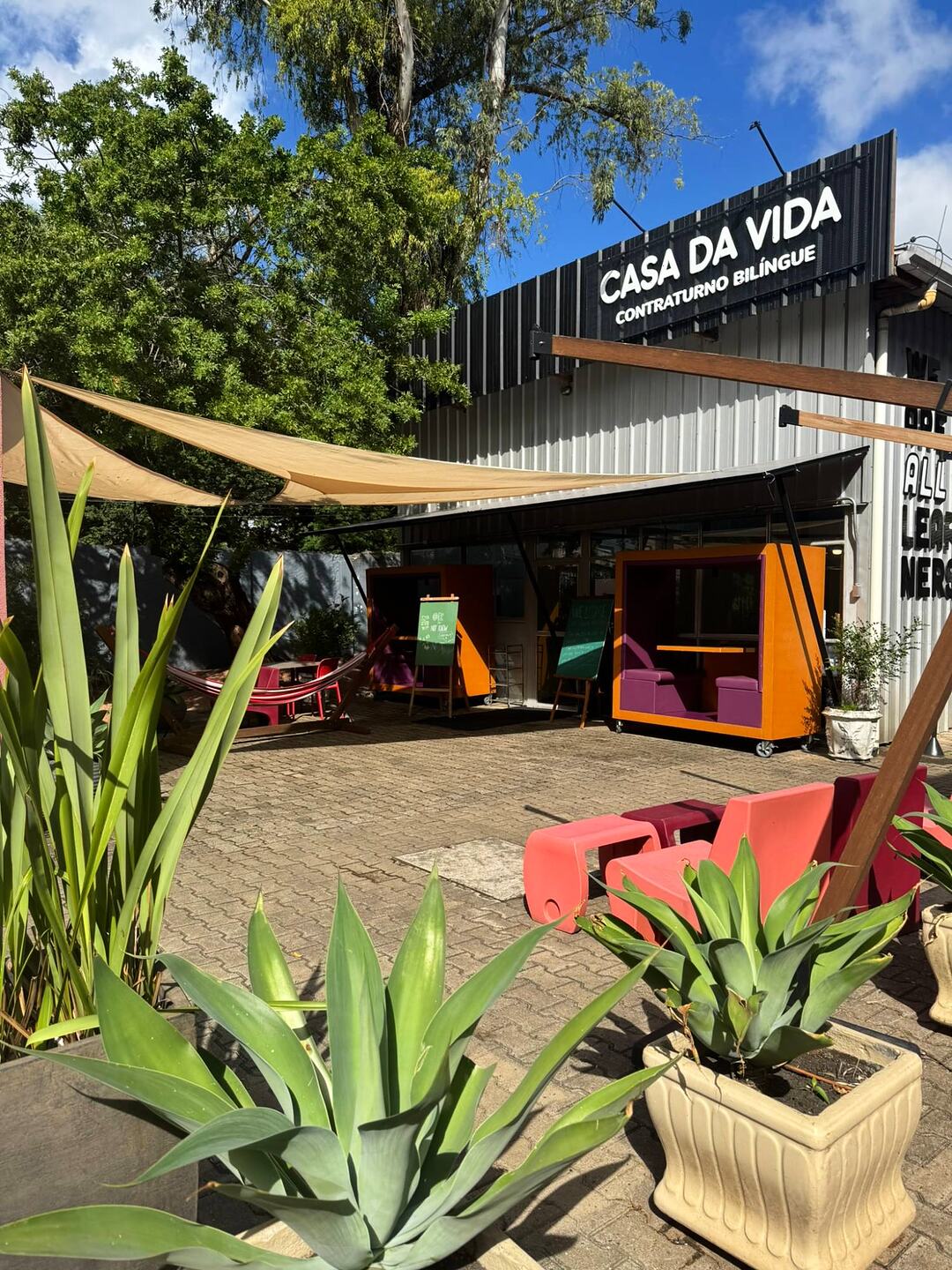 Fachada da Casa da Vida, com a placa Contraturno Bilíngue, cabines coloridas de estudo, rede e plantas no pátio.